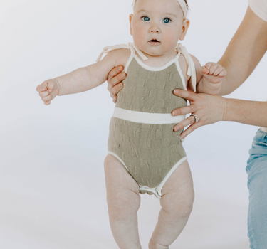 Baby girl wearing organic cotton knit bubble romper in truffle with magnetic opening at crotch and tie shoulder straps.