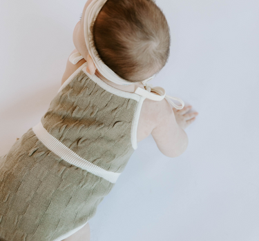 Baby girl wearing organic cotton knit bubble romper in truffle with tie shoulder straps and magnetic closure at crotch.