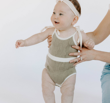 Baby girl wearing organic cotton knit bubble romper in truffle with tie shoulder straps and magnetic closure at crotch.