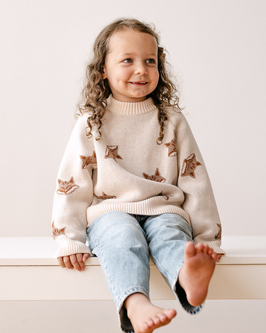 Image shows little girl wearing organic cotton knit sweater with jacquard fox pattern in pale pink. 