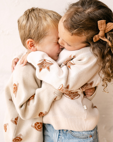 Image shows little girl wearing organic cotton knit sweater with jacquard fox pattern in pale pink and little boy wearing organic cotton knit sweater with jacquard snail pattern in oatmeal. 