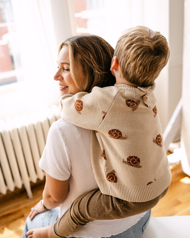 Image shows toddler boy wearing organic cotton knit sweater with snail jacquard pattern in oatmeal. 