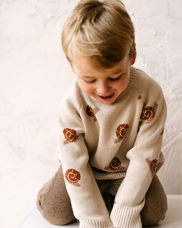 Image shows toddler boy wearing organic cotton knit sweater with snail jacquard pattern in oatmeal. 