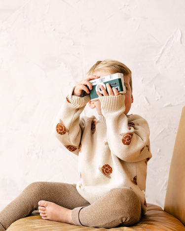 Image shows toddler boy wearing organic cotton knit sweater with snail jacquard pattern in oatmeal. 