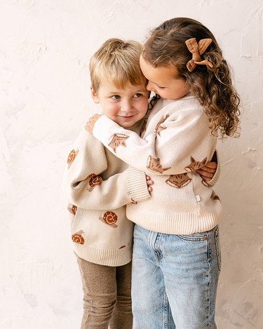 Image shows toddler boy wearing organic cotton knit sweater with snail jacquard pattern in oatmeal. 