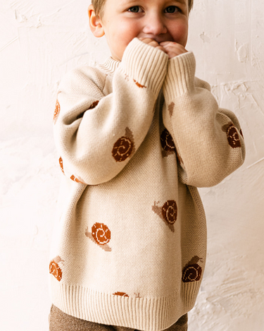 Image shows toddler boy wearing organic cotton knit sweater with snail jacquard pattern in oatmeal. 